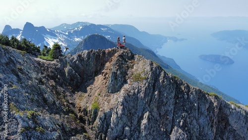 Obraz na płótnie Male adults standing atop a jagged rocky mountain overlooking a tranquil lake below w salonie