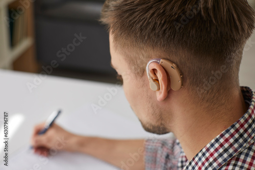 Deaf student with hearing aid. Hearing impaired man, wearing plastic beige hearing device behind ear, is studying at desk at college or at home. Close up of man\'s head. Hearing loss, education concept