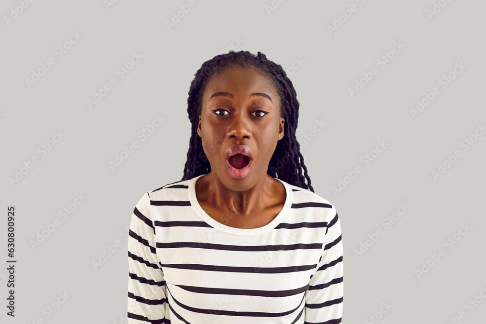 Studio portrait of young African American woman in striped white black long sleeve shirt standing on white background looking at camera with surprised, amazed, happy, open mouthed facial expression