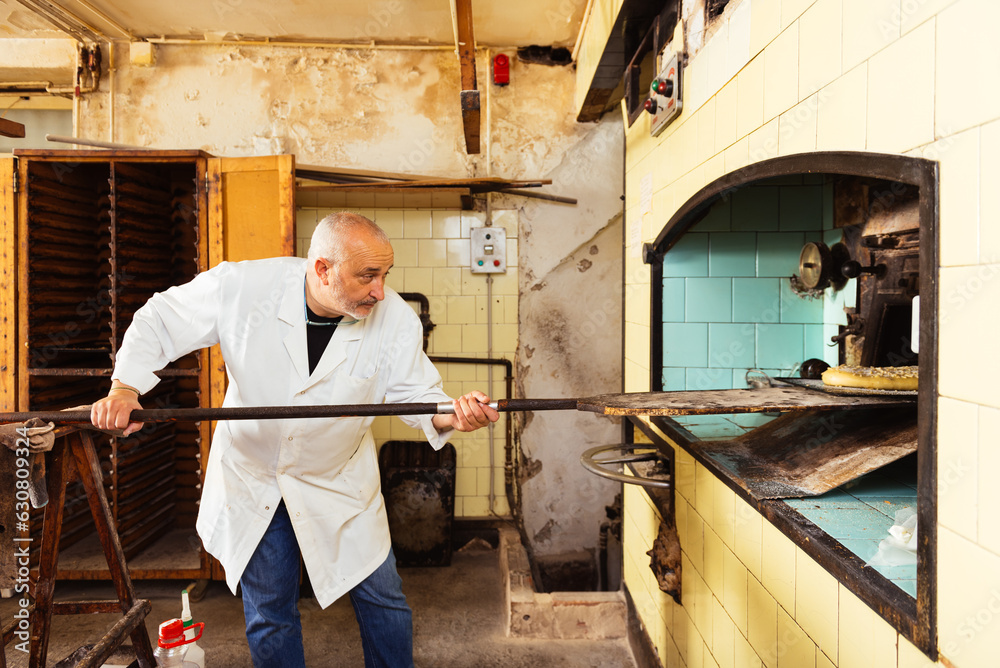 Middle aged baker introducing cake dough into an oven with a wooden shovel Middle aged baker introducing cake dough into an oven with a wooden shovel