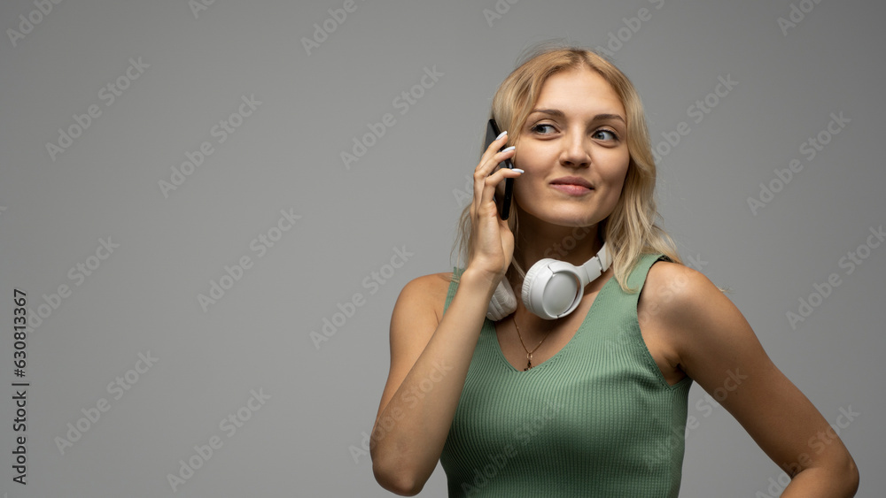Portrait of smiling happy young blonde female woman in green t-shirt and white headphones on a shoulder looking away while talking on mobile phone and standing over black background. Portrait of smiling happy young blonde female woman in green t-shirt and white headphones on a shoulder looking away while talking on mobile phone and standing over black background.