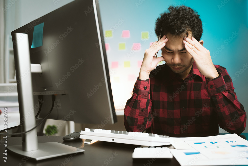 Stressed and tired Asian young man feels headache while working overtime at night in his office. Stressed and tired Asian young man feels headache while working overtime at night in his office.