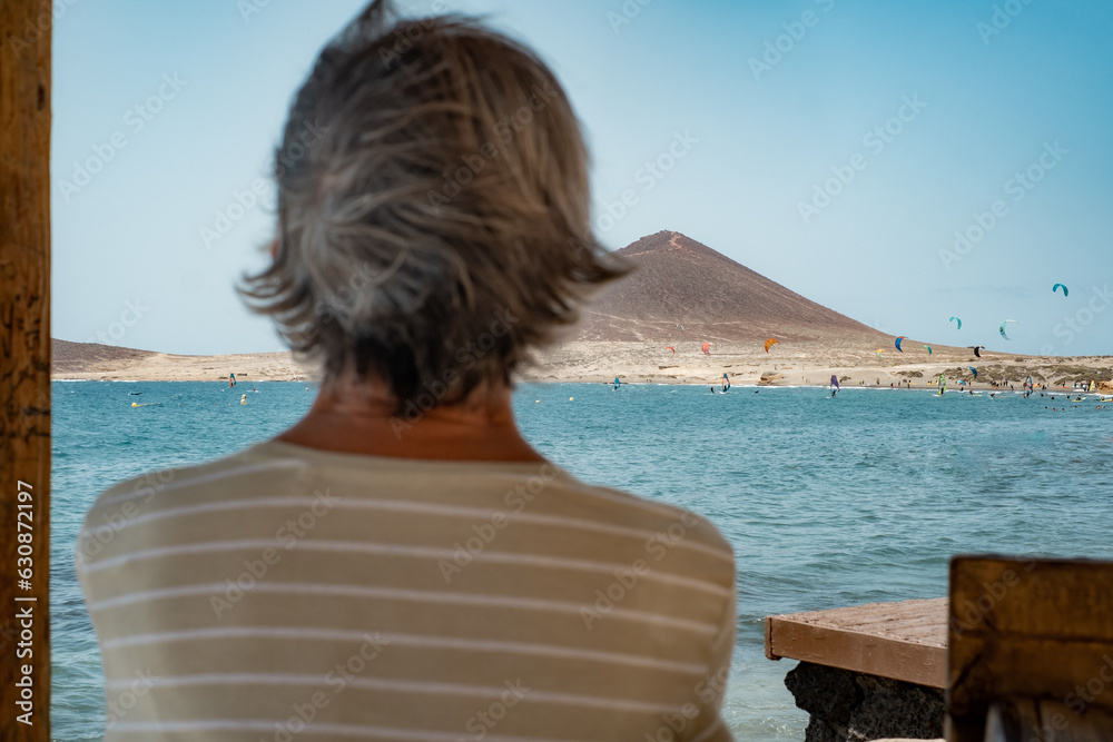 Rear view of mature woman in front of the sea looking at beautiful seascape, alone elderly people enjoying beach vacation or retirement