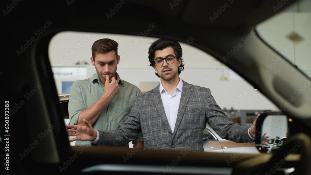 View from interior of car to professional car dealer selling new automobile to pensive male client explaining characteristics. Bearded salesman wearing business suit helping with choice of new auto. View from interior of car to professional car dealer selling new automobile to pensive male client explaining characteristics. Bearded salesman wearing business suit helping with choice of new auto.