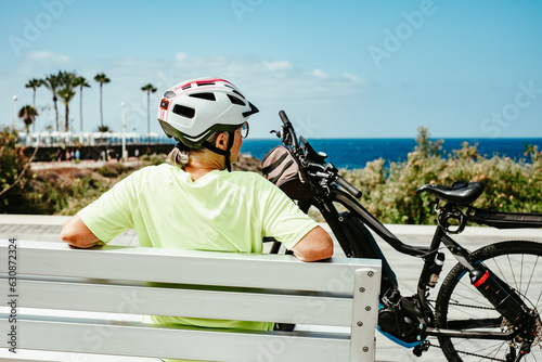 Rear view of senior cyclist woman sitting in a bench close to her bicycle resting looking at the horizon over sea. Elderly carefree lady enjoying freedom, healthy lifestyle and retirement