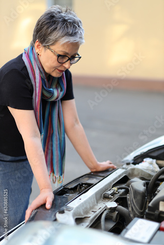 A beautiful and caucasian woman looks at the engine of a car. She is a broken and frustrated driver, having a problem with her vehicle or hood. She is in need of assistance or service.