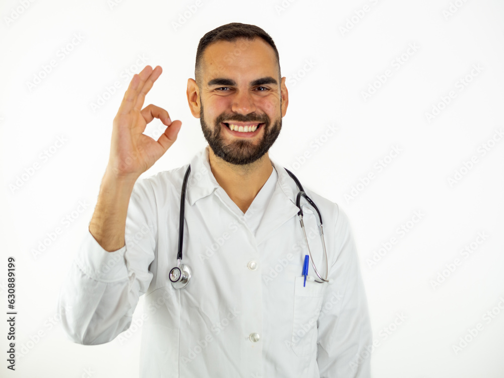 Fototapeta premium Young male doctor ok sign while smiling. Looking at camera isolated on white background