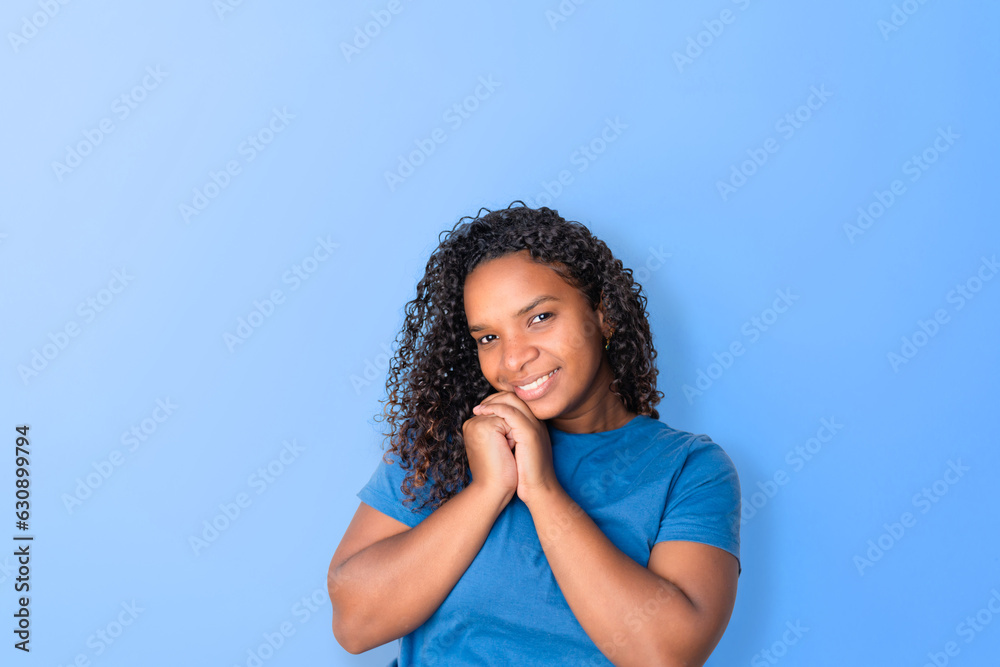 Black woman smiling happily, feeling good and loved, gently clasping hands together, with blue background. Black woman smiling happily, feeling good and loved, gently clasping hands together, with blue background.
