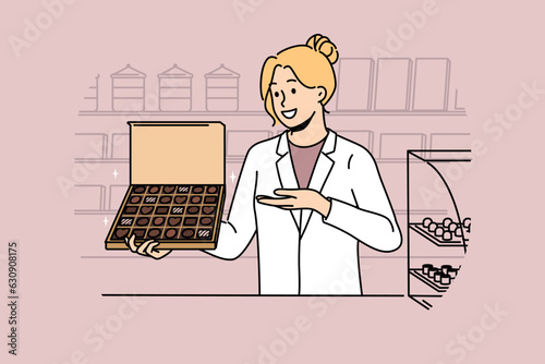 Woman seller with box of chocolates works in confectionery store and offers to buy appetizing dessert made from milk and cocoa. Girl in white coat of seller demonstrates chocolates with smile