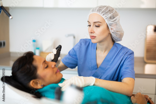 Caucasian woman beautician using equipment for needle-free mesotherapy for her patient  latin woman.