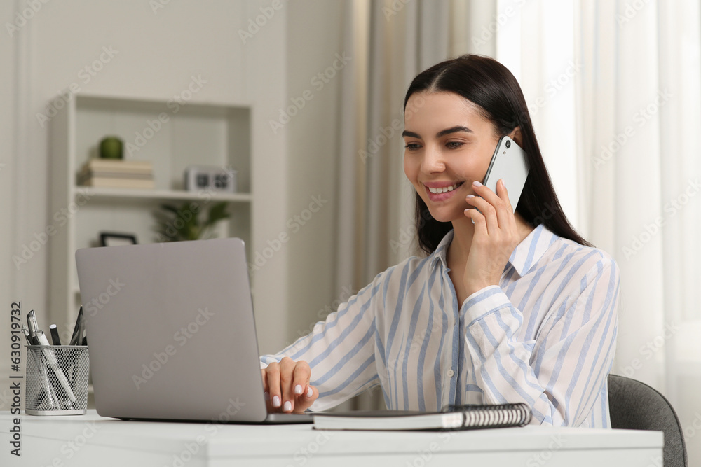 Happy woman talking on smartphone while working with laptop at white desk in room Happy woman talking on smartphone while working with laptop at white desk in room