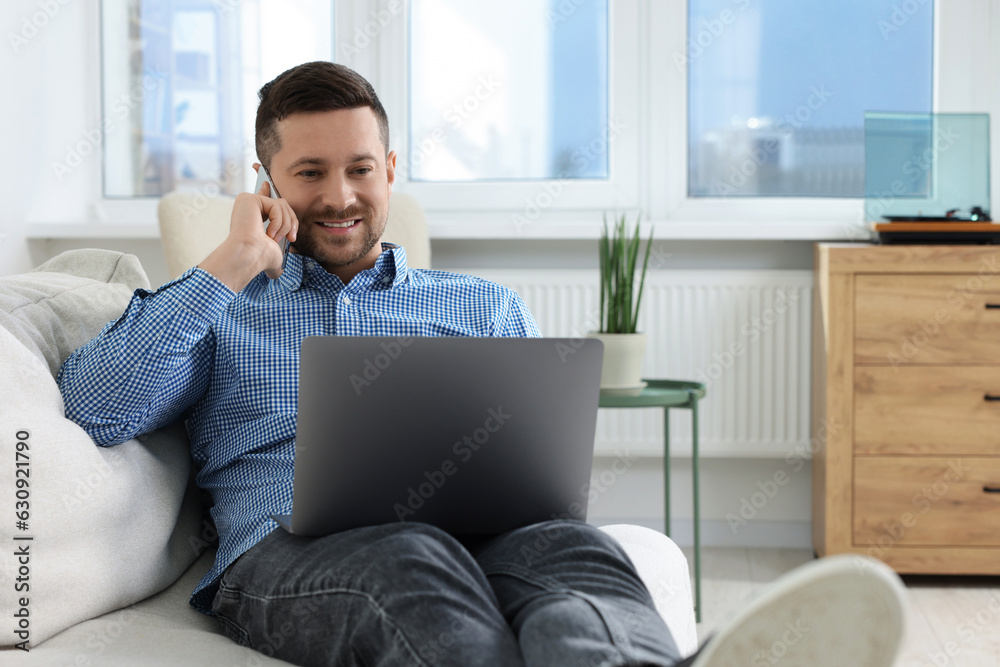 Happy man talking on phone while working with laptop on sofa at home Happy man talking on phone while working with laptop on sofa at home