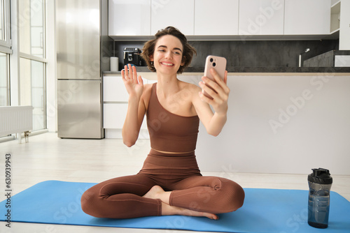 Smiling fitness girl, young athlete sits at home on yoga mat, waves hand at smartphone, joing online workout training session from home, says hello photo