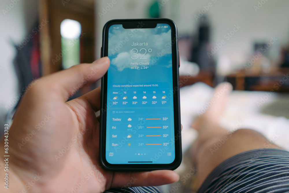 An Asian man holding a smartphone showing weather of Jakarta city, Indonesia while lying on the bed