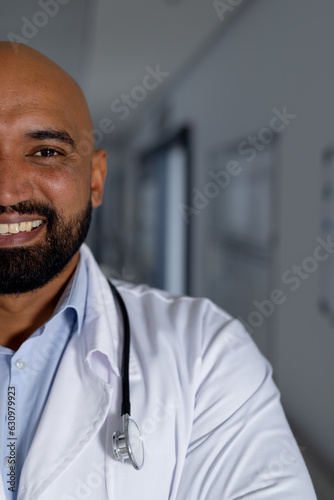 Portrait of happy biracial male doctor wearing lab coat in corridor at hospital, copy space