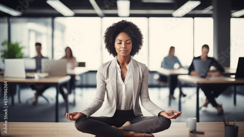 African ethnicity business woman doing yoga in the office sitting on her desk with folded legs and closed eyes, cold lighting
