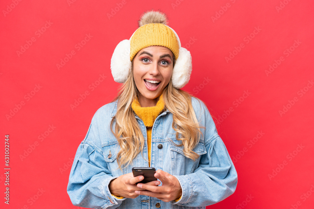 Young pretty Uruguayan woman wearing winter muffs isolated on red background background surprised and sending a message Young pretty Uruguayan woman wearing winter muffs isolated on red background background surprised and sending a message