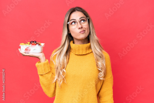 Young pretty Uruguayan woman holding a bowl of fruit isolated on red background and looking up