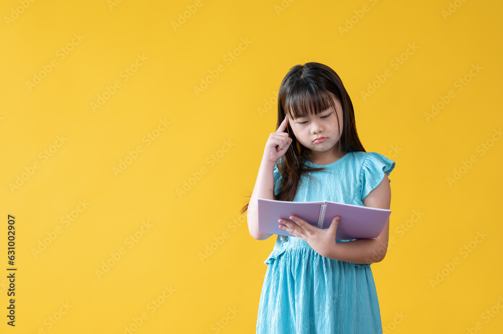 Fototapeta premium An adorable young Asian girl is reading a book, standing against an isolated yellow background