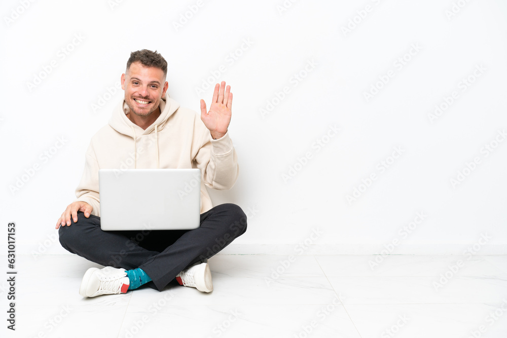 Young caucasian man with a laptop sitting on the floor isolated on white background saluting with hand with happy expression Young caucasian man with a laptop sitting on the floor isolated on white background saluting with hand with happy expression