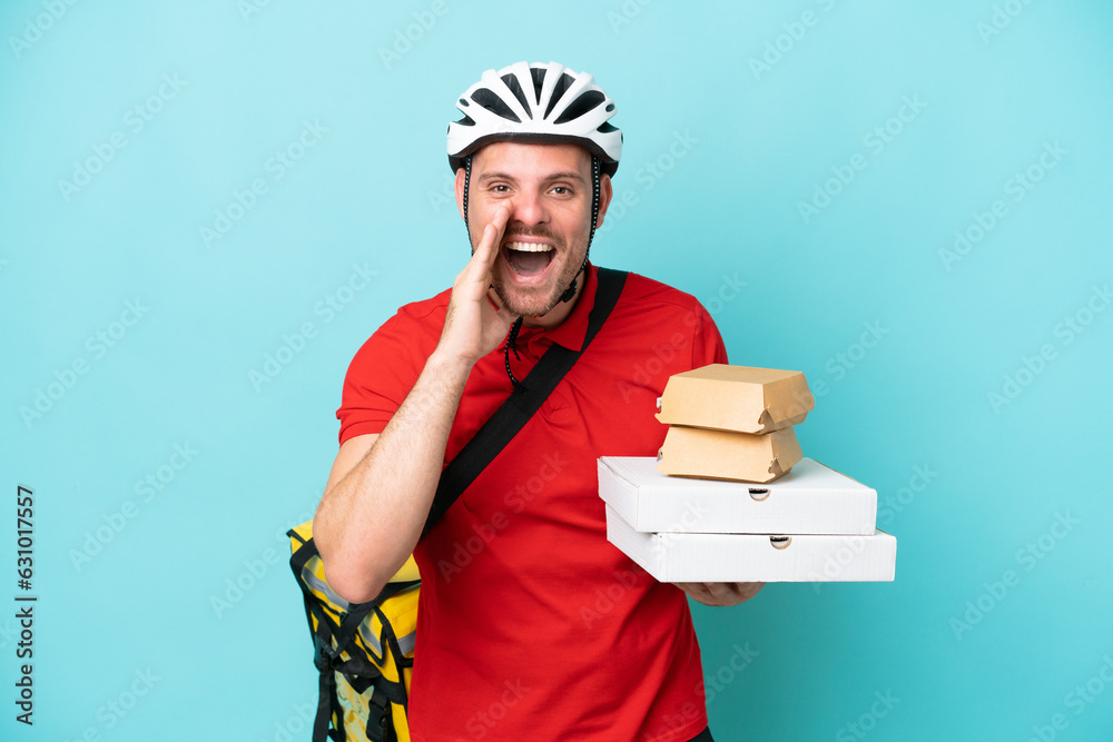 Young caucasian man with thermal backpack and holding fast food isolated on blue background shouting with mouth wide open Young caucasian man with thermal backpack and holding fast food isolated on blue background shouting with mouth wide open