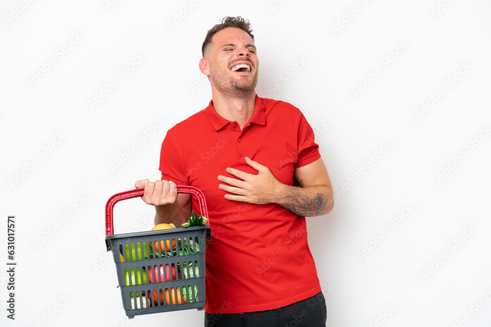 Young caucasian man holding a shopping basket full of food isolated on white background smiling a lot Young caucasian man holding a shopping basket full of food isolated on white background smiling a lot