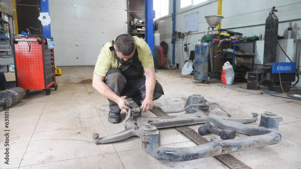Auto mechanic assembles the subframe of car before installation at garage. Professional bearded repairman with special tool fixing automobile parts at workshop. Concept of maintenance and diagnostic Auto mechanic assembles the subframe of car before installation at garage. Professional bearded repairman with special tool fixing automobile parts at workshop. Concept of maintenance and diagnostic