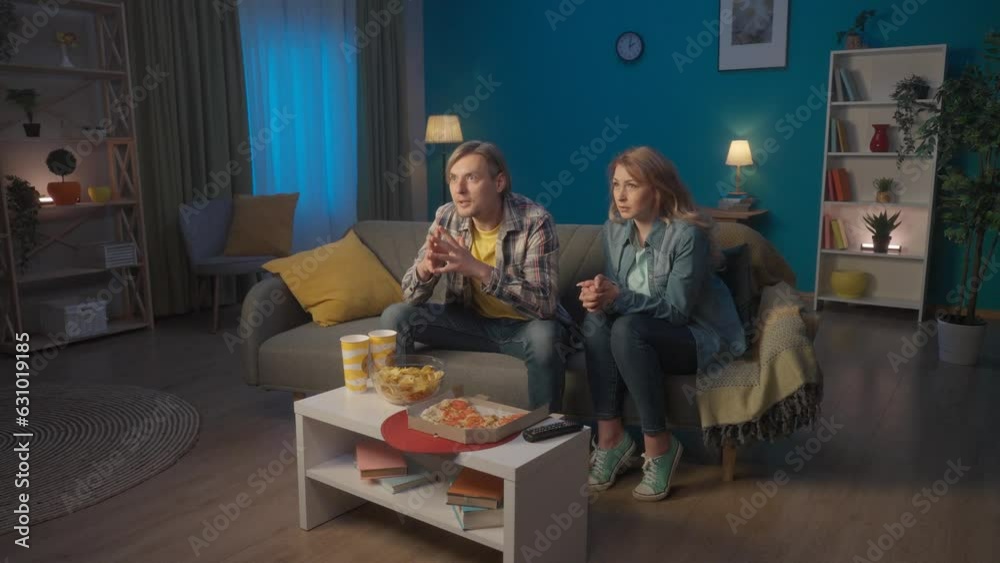 A couple watches a sports channel on TV in the evening at home. A man watches the game, gets upset during a loss, a woman cheers and reassures him. Slow motion. HDR BT2020 HLG Material. A couple watches a sports channel on TV in the evening at home. A man watches the game, gets upset during a loss, a woman cheers and reassures him. Slow motion. HDR BT2020 HLG Material.