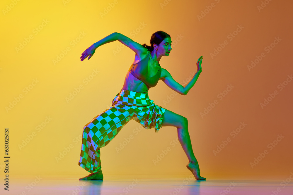 Artistic, dynamic image of young woman in motion, training, dancing against gradient yellow orange background in neon light. Concept of modern dance style, hobby, art, performance, lifestyle, ad