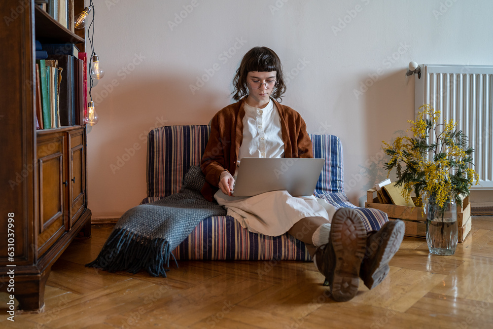 Focused freelancer girl sit on couch with laptop crossed legs in boots at home writes essay, draws project, searches information on internet webs. Interested business woman working in creative startup Focused freelancer girl sit on couch with laptop crossed legs in boots at home writes essay, draws project, searches information on internet webs. Interested business woman working in creative startup