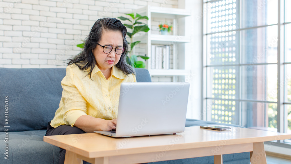 Asian old senior pensioner chubby fat housewife sitting on cozy sofa  while surfing browsing internet online via laptop computer in living room.