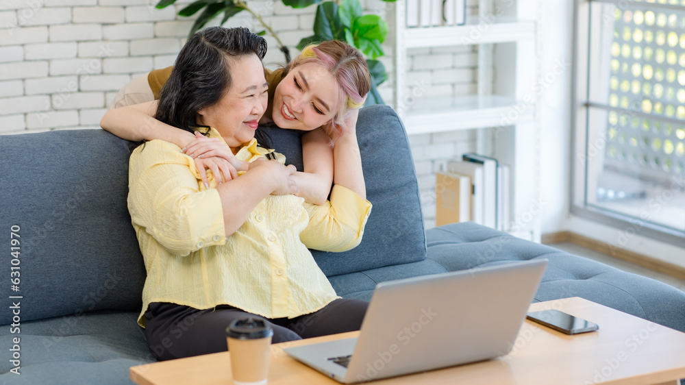 Asian young cheerful pretty female daughter standing behind old chubby senior retirement pensioner mother sitting on sofa in living room at home holding hands hugging cuddling showing love together Asian young cheerful pretty female daughter standing behind old chubby senior retirement pensioner mother sitting on sofa in living room at home holding hands hugging cuddling showing love together