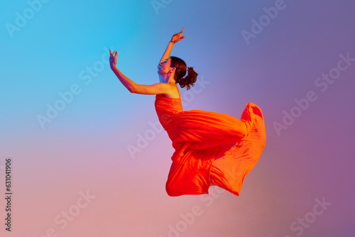 Dynamic image of artistic young woman dancing in elegant red dress against gradient multicolor background in neon light. Concept of modern dance style, hobby, art, performance, lifestyle, ad photo