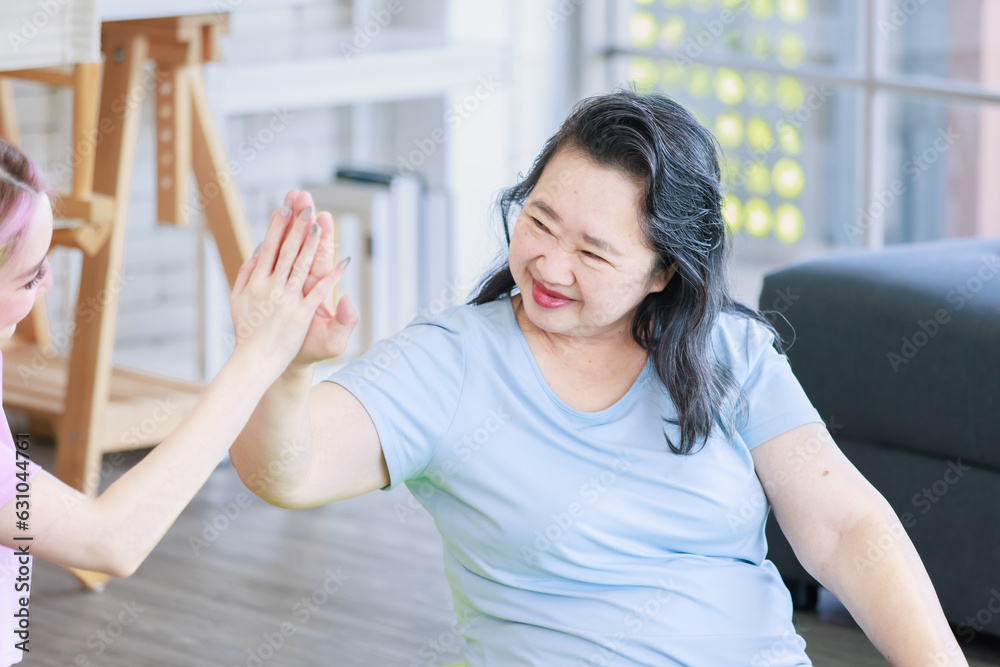 Fototapeta premium Asian old senior pensioner retirement cheerful happy healthy fit mother in casual sportswear and young beautiful daughter holding raising hand high five watching doing yoga stretching in living room.
