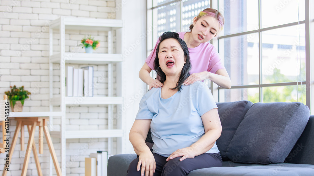Asian young beautiful daughter standing behind massaging neck of old senior pensioner having backache from stretching in casual sportswear sitting on sofa couch in living room at home. Asian young beautiful daughter standing behind massaging neck of old senior pensioner having backache from stretching in casual sportswear sitting on sofa couch in living room at home.