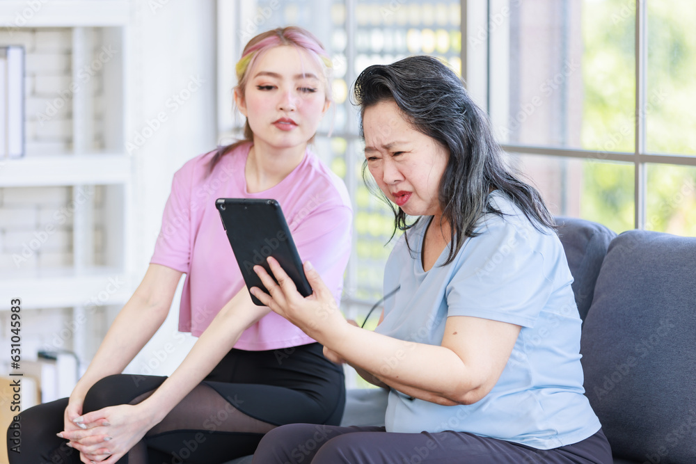 Asian young beautiful daughter and old mother senior pensioner retirement try to see tablet computer sitting on sofa couch in living room at home Asian young beautiful daughter and old mother senior pensioner retirement try to see tablet computer sitting on sofa couch in living room at home