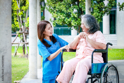 Asian female doctor in surgical gown caring for an elderly female patient sitting in a wheelchair outside a building. medical services in hospitals