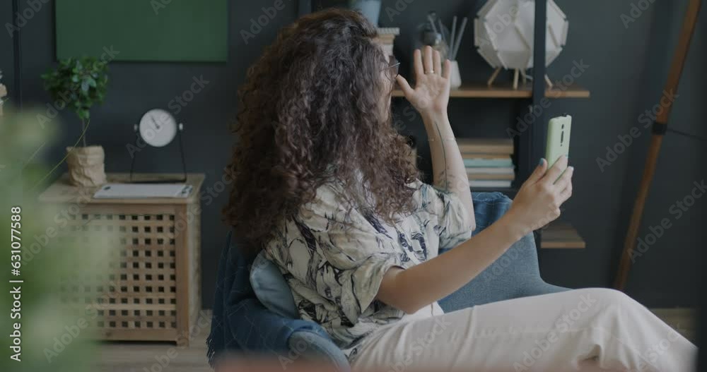 Portrait of joyful young woman making online video call using smartphone talking waving hand sending air kiss in armchair at home. People and communication concept. Portrait of joyful young woman making online video call using smartphone talking waving hand sending air kiss in armchair at home. People and communication concept.