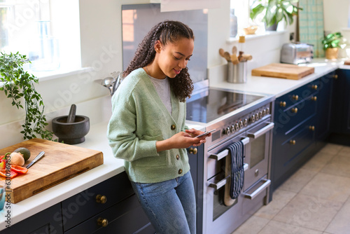 Teenage Girl At Home In Kitchen Connecting With Friends On Social Media Using Mobile Phone