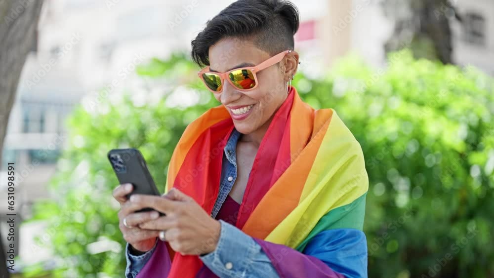 Young beautiful hispanic woman using smartphone wearing rainbow flag at park Young beautiful hispanic woman using smartphone wearing rainbow flag at park