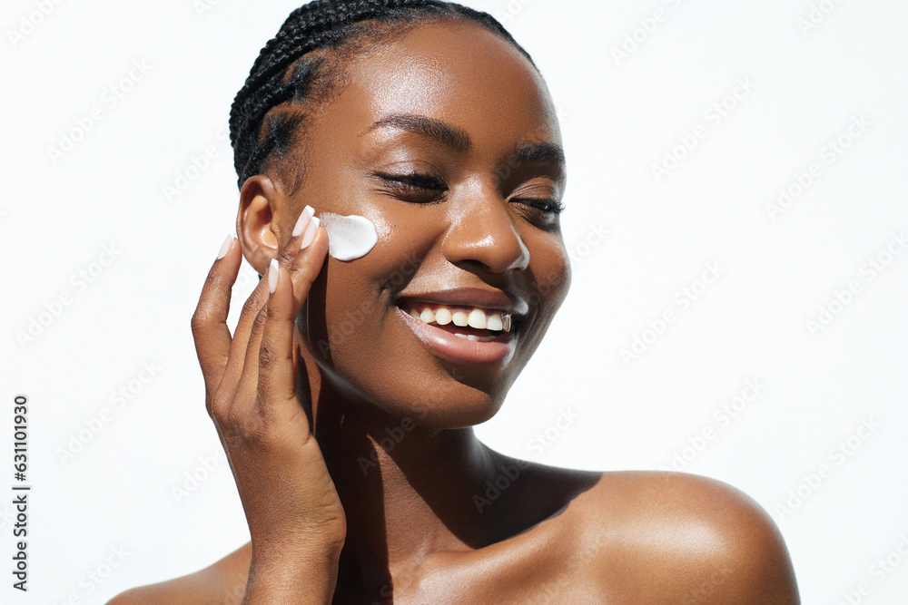 Beauty and skincare. Skin hydration and oil balance. Portrait of African American young woman with afro braids hairstyle is applying a cream smear on her face and standing against white background - obrazy, fototapety, plakaty 