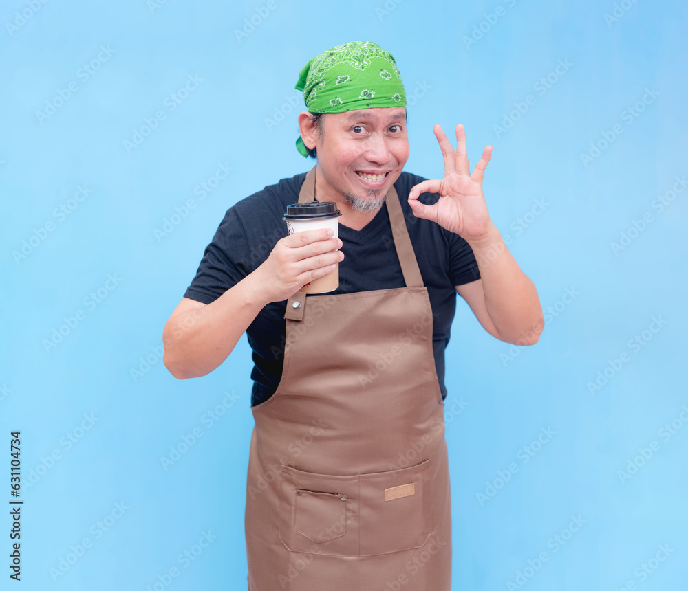 A happy middle-aged barista holding a cup of coffee to-go on one hand while doing an okay sign. Isolated on a blue background.