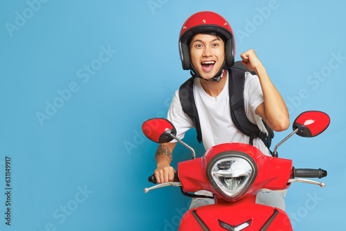 Asian happy guy in red helmet riding red motorbike and holds one fist up, posing on blue backdrop with wide smile, lifestyle concept, copy space