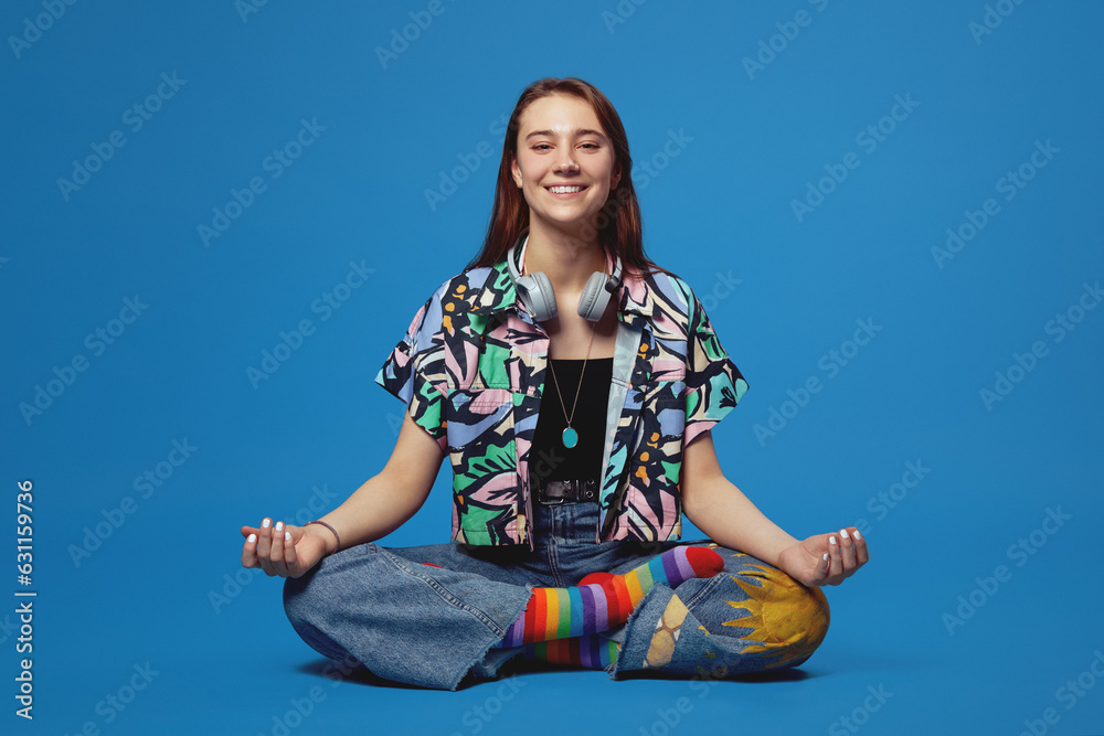 Photo of full body pretty young focused girl smiling and meditating, isolated over blue background. Cute bright female with headphones looking at camera while sitting on floor in lotus pose. Photo of full body pretty young focused girl smiling and meditating, isolated over blue background. Cute bright female with headphones looking at camera while sitting on floor in lotus pose.