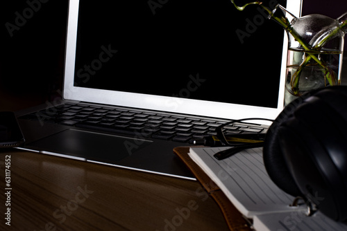 Workplace with open laptop, a blank open note book, a smart phone ,a pen, a specs and a head phone on dark wooden desk