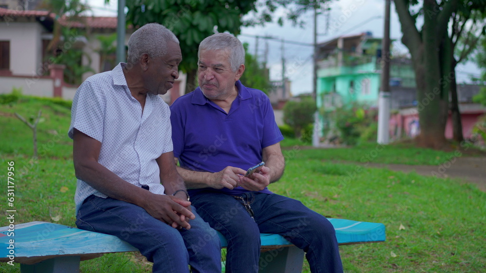 Two diverse seniors looking at phone sitting on park bench outdoors laughing and smiling together. Older friend sharing online media content to companion depicting authentic friendship Two diverse seniors looking at phone sitting on park bench outdoors laughing and smiling together. Older friend sharing online media content to companion depicting authentic friendship