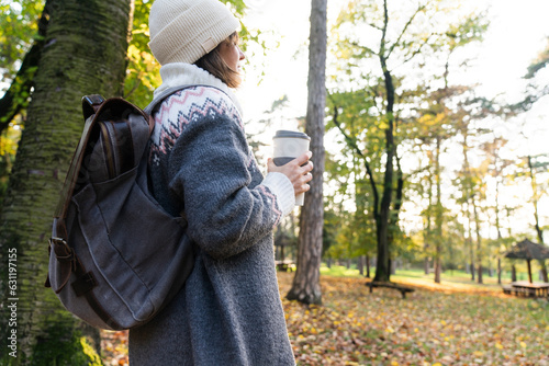 Woman traveler with backpack is holding a cup of hot drink in autumn park..