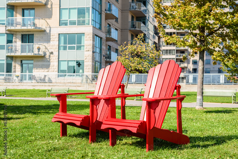 Red Adirondack chairs on a grass in a park o na sunny autumn day. Apartment buildings are visible in background.