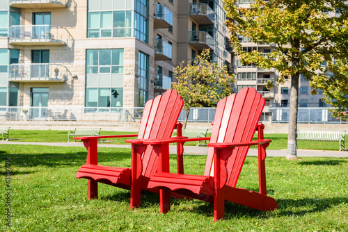 Red Adirondack chairs on a grass in a park o na sunny autumn day. Apartment buildings are visible in background. © alpegor