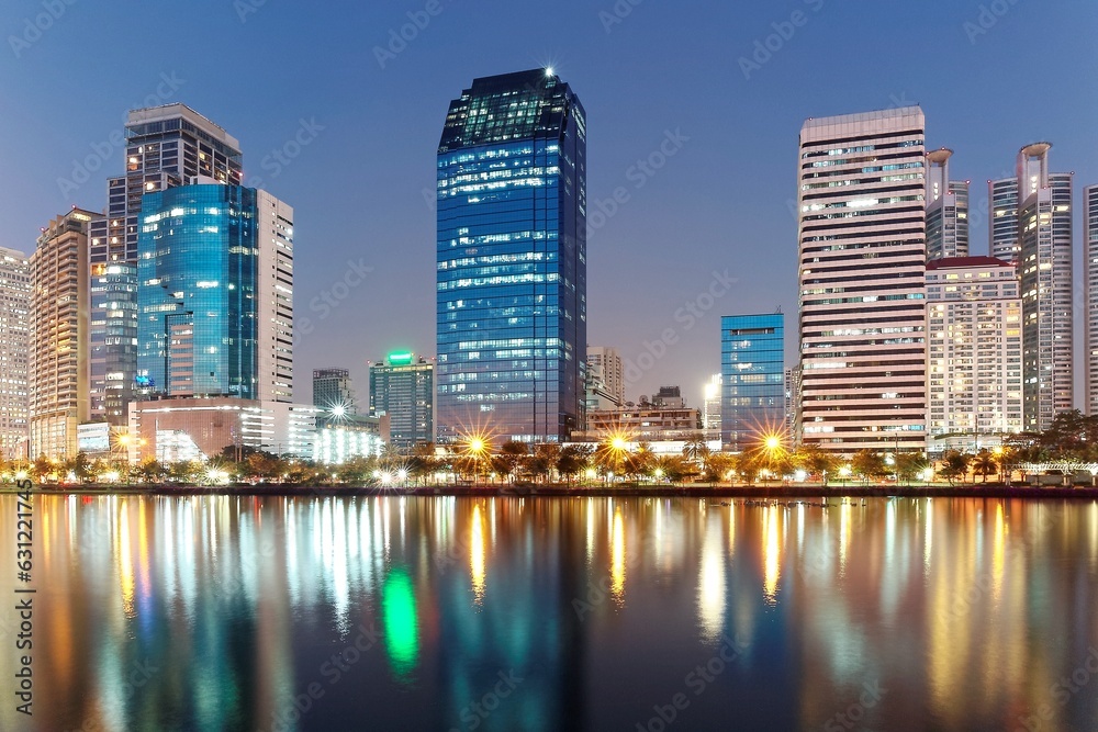 Night skyline of modern skyscrapers by lakeside, with glass curtain walls and dazzling city lights reflected in the smooth lake water in beautiful Benjakiti Park at blue dusk, in Bangkok, Thailand